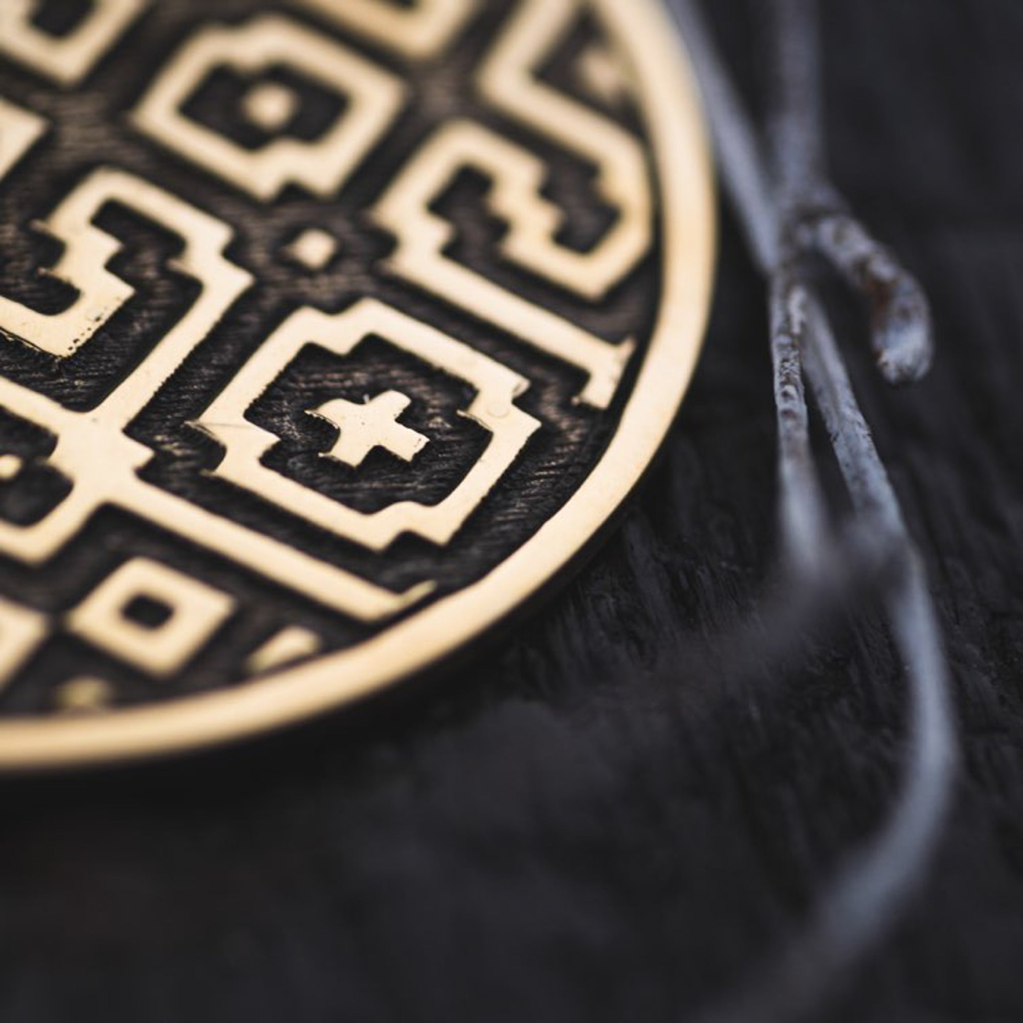 A close-up photo of a round, golden earring with an intricate Sacred Geometry Shipibo pattern from the AREVALO Shipibo Oversized Disc Hoop Earrings in Gold by Kaya Movement. The motif includes squares, crosses, and other shapes set against a dark, textured background, with two thin metallic wires partially visible on the right side of the image.